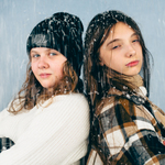 Dwie dziewczyny opierają się plecami o siebie, jedna jest w czapce. Z góry sypie na nie śnieg.