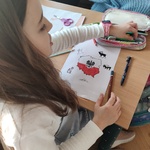 Dziewczynka rysuje plakat na ławce. Jedną ręką bierze do ręki kredkę z piórnika.