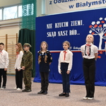 Grupa uczniów ubranych w stroje galowe występuje na uroczystej akademii. W tle flaga Polski oraz dekoracja patriotyczna na niebieskim płótnie.